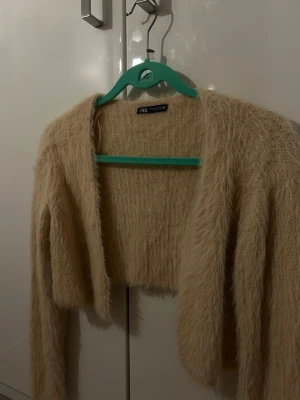 croppad kofta från Zara - Supermjuk och fluffig beige kofta från Zara. Croppad modell med lång ärm och öppen framtill. Perfekt för lager på lager och ger en cozy vibe till din outfit. Materialet är stickat med en lätt glittrig finish som syns i ljuset.