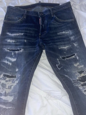 Dsquared2 blå slitna jeans, storlek 48 - Säljer ett par blå jeans från Dsquared2 i storlek 48. Jeansen har en skinny passform med flera slitningar och trasiga detaljer framtill för en riktigt edgy look. Klassisk femficksmodell och tillverkade i jeansmaterial. Perfekta för dig som gillar streetwear och statement-plagg.