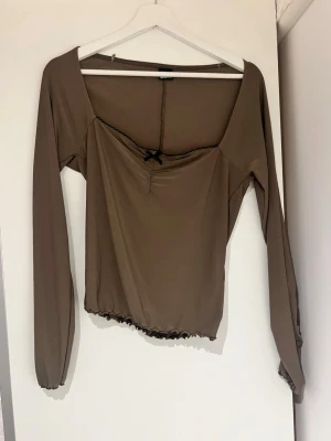 Brun långärmad topp med rosett - Säljer en brun långärmad topp med fyrkantig urringning och liten svart rosett framtill. Toppen har rynkade detaljer vid bysten och svarta vågiga kanter nedtill och vid ärmsluten. Perfekt att styla med jeans eller kjol.