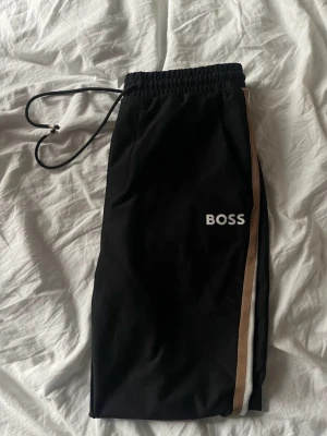 ***Svarta mjukisbyxor från BOSS*** - Svarta mjukisbyxor från BOSS med beige och vit sidoremsa. Byxorna har resår i midjan och snörning för justerbar passform. 