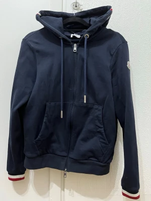 Mörkblå hoodie från Moncler - Snygg mörkblå hoodie från Moncler med dragkedja, stor huva och dragsko. Klassisk känguruficka framtill och Moncler-logga på ärmen. Ribbade muddar med rödvita detaljer ger en sportig vibe. Perfekt för dig som vill ha både stil och komfort.