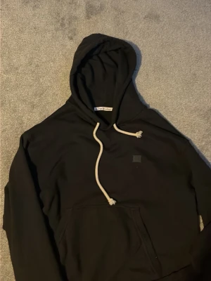 Svart hoodie från Acne Studios - Säljer en svart hoodie från Acne Studios med vit dragsko och klassisk känguruficka framtill. Hoodien har en liten diskret logopatch på bröstet och är tillverkad i mjukt bomullsmaterial. Perfekt för en avslappnad och stilren look.