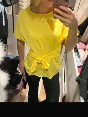 Gul topp med knyt från Pieces - Säljer en solgul topp från Pieces med rund halsringning och korta ärmar. Toppen har ett brett knytband i midjan som ger en snygg siluett och kan stylas på olika sätt. 