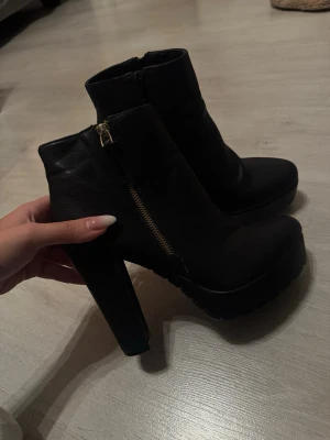Svarta stövletter med platå och klack - Använda, en gång inomshus! Säljer ett par svarta stövletter i skinnimitation med chunky hög klack och platåsula. Skorna har dragkedja på sidan i guld och rund tå. Perfekta för dig som vill ha lite extra höjd och edgy stil. 