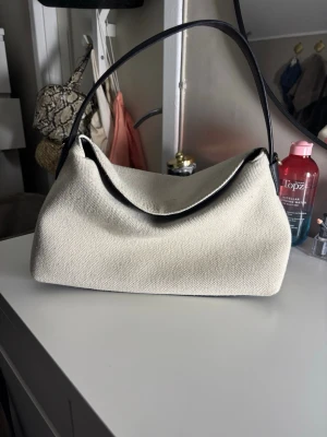 Beige axelväska i canvas - Stilren beige axelväska i canvas med svart axelrem och ljus insida. Väskan har en enkel, rektangulär form och är perfekt för dig som gillar minimalistisk design. Insidan är fodrad i ljus textil och har en liten innerficka för småsaker.