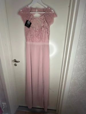 Rosa spetsklänning från Nelly  - Lång rosa klänning från Nelly med spets upptill och vackra detaljer. Klänningen har korta ärmar, öppen rygg och ett knytband i midjan. Perfekt för speciella tillfällen när du vill känna dig extra fin.