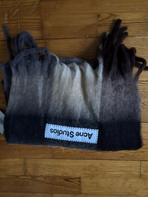 Gråmelerad halsduk från Acne Studios - Säljer en fluffig och mjuk halsduk från Acne Studios i gråmelerade toner med snygg ombre-effekt. Halsduken har långa fransar i ändarna och en tydlig logotyp-patch. Perfekt accessoar för att lyfta din outfit.