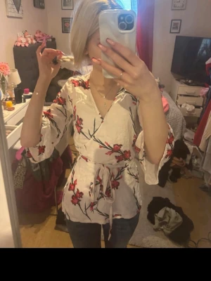 Vit omlottblus med blommigt mönster - Snygg vit omlottblus med rosa och röda blommor samt svarta grenar. Blusen har trekvartsärmar med volang och knytskärp i midjan för en feminin siluett. Perfekt för att lyfta din outfit med en romantisk touch.