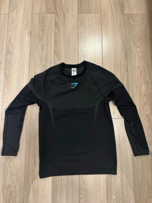 Onyx GYMSHARK  - Svart långärmad träningströja från Gymshark med diskreta mönsterdetaljer på axlar och ärmar. Tröjan har rund halsringning och blå Gymshark-logga på bröstet. Tillverkad i ett stretchigt funktionsmaterial som andas, perfekt för träning. Helt oanvänd utan prislapp.