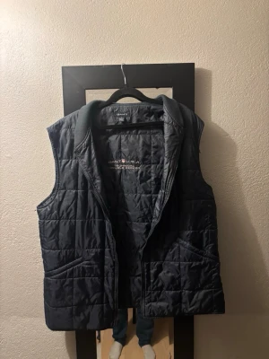 Mörkblå dunväst från Gant - Snygg mörkblå dunväst från Gant med quiltat mönster och ribbad krage. Västen har två fickor framtill och dragkedja. Perfekt för lager på lager och ger en sportig look. Märkt med 'GANT U.S.A. Technical Double Blocker' på insidan.