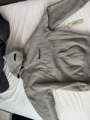 Grå Essentials Fear of God hoodie - Säljer en grå hoodie från Essentials Fear of God med svart logga tryckt på bröstet och ryggen.  Pris kan diskuteras 
