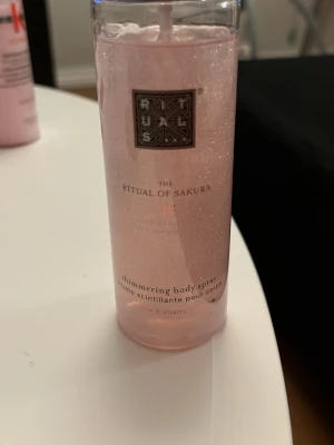 Rituals  Body Spray - Shimmering body spray från Rituals, . Flaskan är genomskinlig med rosa skimmer.  doft av rismjölk och körsbärsblom. Perfekt för dig som vill ha en glittrande finish på huden.