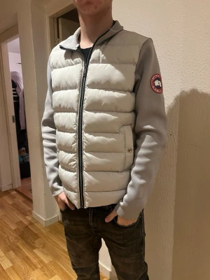 Ljusgrå cardigan från Canada Goose - Snygg ljusgrå cardigan från Canada Goose med quiltad front och dragkedja. Jackan har två fickor med knappstängning och klassisk logga på ärmen. Perfekt för lager-på-lager och riktigt clean look. Passar S/M. Killen på bild är 1.78 och S. Använd gärna köp nu