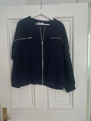 Mörkblå bomberjacka från Wera - Säljer en mörkblå bomberjacka från Wera med silverfärgade dragkedjor framtill och på bröstet. Jackan har en loose passform, långa ärmar och praktiska fickor på sidorna. Materialet är mjukt och känns skönt mot huden. Perfekt för dig som gillar en clean och stilren look.