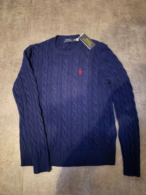 Marinblå kabelstickad tröja Polo Ralph Lauren - Kabelstickad marinblå tröja från Polo Ralph Lauren med rund halsringning och röd broderad logga på bröstet. Tröjan har ribbade muddar vid ärmslut och nederkant. Klassisk och tidlös design som passar till mycket.