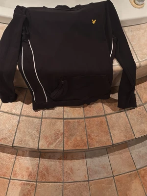 Svart hoodie från Lyle & Scott - Svart hoodie från Lyle & Scott med vit piping längs sidorna och klassisk gul logga på bröstet. Har huva och magficka framtill. Perfekt för en chill och sportig look.