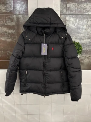 Svart pufferjacka från Polo Ralph Lauren - Snygg svart pufferjacka från Polo Ralph Lauren med huva och dragkedja framtill. Jackan har två sidofickor med dragkedja och den klassiska röda loggan broderad på bröstet. Perfekt för kalla dagar och riktigt trendig look.