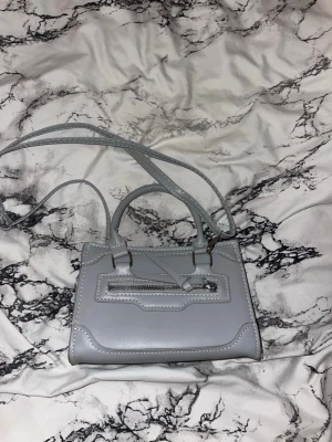 Väska - Väska från Shein, går att hålla som handväska annars ett justerbart band som går över kroppen/axeln. Aldrig använd. Färgen är grå/blå, skulle själv anse den som grå. 