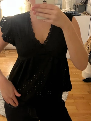 Svart blus med spets och hålmönster - Säljer en svart blus med v-ringning och spetskant. Blusen har korta ärmar med volang och ett fint hålmönster över hela tyget. Passformen är lite loose och toppen ger en söt och trendig look.