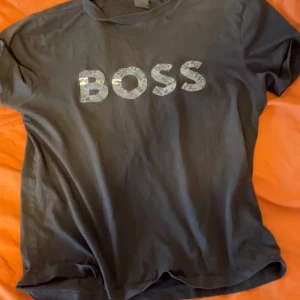 Svart BOSS t-shirt med logga - Snygg svart t-shirt från BOSS med stor logga framtill i grått och vitt mönster. Klassisk rund hals och korta ärmar. Perfekt till jeans eller shorts för en clean och enkel stil.