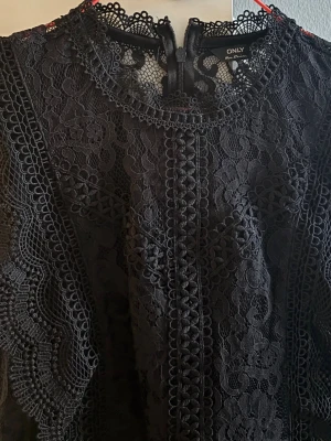 Svart spetsblus från ONLY - Supersnygg svart blus från ONLY i spets med fina detaljer och rund halsringning. Ärmlös modell med vackra spetskanter och knäppning bak i nacken. Perfekt att styla med jeans eller kjol för en trendig look.