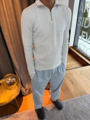 Vit ribbad tröja med halv dragkedja - Snygg vit långärmad tröja med ribbad struktur och halv dragkedja vid halsen. Tröjan har en smal passform och en liten krage som ger en clean och modern look. Perfekt att matcha med jeans eller chinos för en stilren outfit.