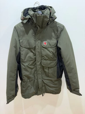 Grön dunjacka från Fjällräven - Snygg grön dunjacka från Fjällräven med svarta detaljer på sidor och ärmar. Jackan har huva, flera praktiska fickor och dragkedja framtill. Perfekt för kalla dagar och outdoor-äventyr. Klassisk Fjällräven-logo på bröstet.