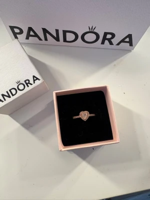 Pandora ring med hjärtformad sten - Säljer en snygg ring från Pandora i roséguld med en rosa hjärtformad sten. Säljer då den ej kommer till användning. Storlek 54 som motsvarar 17.20 i mm