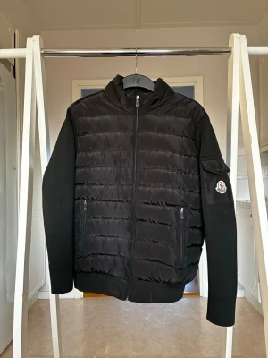 Svart pufferjacka från Moncler - Säljer en svart pufferjacka från Moncler med quiltad front och rygg, samt stickade ärmar. Jackan har dragkedja framtill, två fickor med dragkedja och en ficka med lock på ärmen där Moncler-loggan sitter. Perfekt för dig som vill ha en stilren och sportig look.