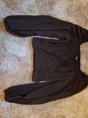 Svart croppad topp med puffärm - Trendig svart croppad topp från H&M med långa puffärmar och smockad rygg. Toppen har en snygg v-ringning och knyt framtill som ger en extra detalj. Perfekt att styla med höga jeans eller kjol för en cool look. 