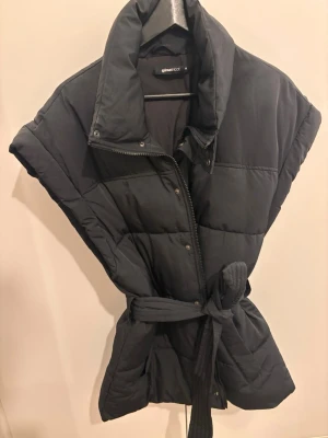 Svart dunväst från Gina Tricot - Snygg svart dunväst från Gina Tricot med hög krage, dragkedja och tryckknappar framtill. Västen har ett bälte i midjan för en mer markerad siluett och är quiltad för extra värme. Perfekt att slänga över en hoodie eller tröja när det är kyligt ute.