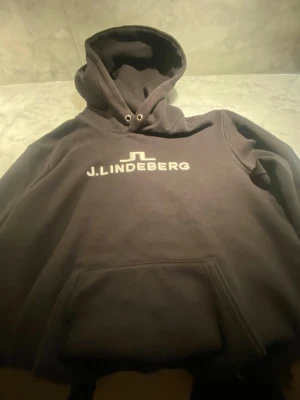 Svart hoodie från J.Lindeberg - Snygg svart hoodie från J.Lindeberg med stor logga broderad i vitt på bröstet. Klassisk modell med huva och känguruficka framtill. Tillverkad i mjukt bomullsmaterial som känns skönt mot huden. Perfekt för en avslappnad och trendig look.
