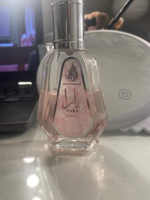 Yara parfym 50ml Ard Al Zaafaran - Elegant parfymflaska med ljusrosa vätska från Ard Al Zaafaran, modellen Yara. Flaskan har en unik, multifacetterad form med silverfärgat lock och dekorativ etikett framtill. 