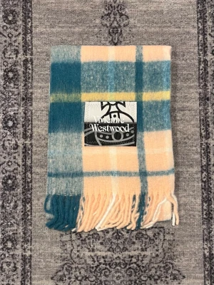 Rutig halsduk från Vivienne Westwood - Säljer en snygg rutig halsduk från Vivienne Westwood i mjuk ullblandning. Färgerna är blågrön, beige, gul och vit med klassiskt rutmönster och fransar i ändarna. Perfekt accessoar för att lyfta din outfit med en cool touch.
