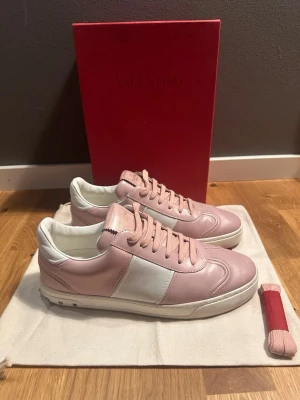 Valentino Open rosa och vita sneakers - Snygga Valentino Open sneakers i ljusrosa och vitt skinn med klassisk låg siluett. Skorna har vita paneler på sidorna, rosa snörning och diskreta nitar vid hälen. Perfekta för dig som vill ha en stilren och trendig look.