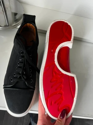 Christian Louboutin svarta sneakers - Säljer ett par svarta sneakers från Christian Louboutin med klassisk röd sula och vita kanter. Skorna har snörning, rund tå och är tillverkade i skinn med beige innersula. Riktigt snygga och sticker ut med den ikoniska röda undersidan. Köpta på vinted men vart för stora, vet ej om dem är äkta, därav priset.