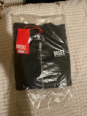 Svart hoodie med dragkedja från Diesel. 12 åring  - Svart hoodie från Diesel med vit logga på bröstet och hel dragkedja framtill. Klassisk huva och fickor, tillverkad i mjuk bomull. Snygg och enkel design med röda tags kvar. Perfekt för en chill och stilren look.