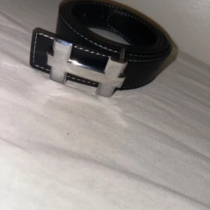 Svart bälte med H-spänne - Snyggt svart bälte i läder med ett stort, silvrigt H-format metallspänne. Perfekt accessoar för att lyfta vilken outfit som helst och ger en clean och modern look. Passar både till jeans och kostymbyxor.