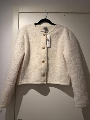 Krämvit bouclékofta från Gina Tricot - Stilren krämvit bouclékofta från Gina Tricot med rund halsringning och dekorativa guldknappar framtill. Helt oanvänd