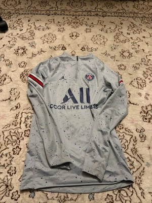 Grå PSG långärmad träningströja - Snygg grå långärmad träningströja från PSG och Jordan med mörkblå logga och text. Tröjan har diskreta svarta prickar, röd-vita ränder på ärmarna och dragkedja vid halsen. Perfekt för fotbollsträning eller chill. Materialet är funktionspolyester. Jag har vanligtvis på mig S, den här som xs passar mig jättebra 