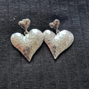 Silverfärgade hjärtformade örhängen - Säljer ett par coola örhängen i silverfärg med hamrad yta. De har dubbla hjärtan, ett mindre upptill och ett större nedtill, vilket ger en unik och charmig look. Perfekt för dig som vill sticka ut med ett statement-smycke.