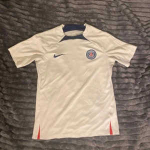 PSG fotbollströja  - Snygg vit PSG fotbollströja från Nike i slim fit-modell. Tröjan har korta ärmar, mörkblå Nike-logga och PSG-emblem på bröstet samt mörkblå detaljer vid halsen. Tillverkad i lätt och funktionellt material, perfekt för träning eller match.