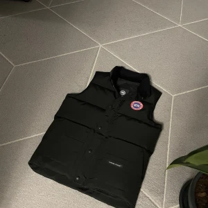 Svart dunväst från Canada Goose - Svart dunväst från Canada Goose med klassisk röd och vit logotyp på bröstet. Västen har hög krage, tryckknappar och två stora fickor framtill. Materialet är slitstark polyester och dunfyllning som håller dig varm. Perfekt för kalla dagar och snygg att styla över hoodie.