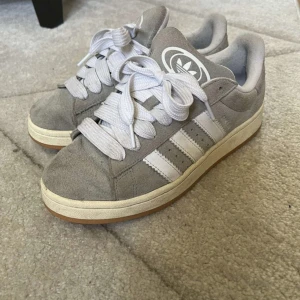 Grå Adidas Campus sneakers  - Säljer ett par grå Adidas Campus sneakers i mocka med vita detaljer och klassiska tre ränder på sidan. De är i bra skick knappt använda💕