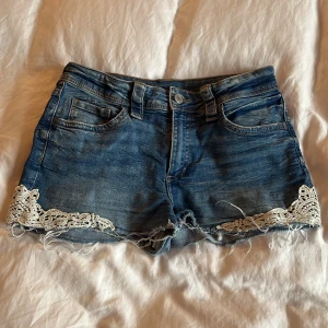 Blå jeansshorts med spets från H&M - Snygga blå jeansshorts från H&M med råa kanter och vit spetsdetalj längs bensluten. Klassisk femficksmodell med bälteshällor och knappstängning. Perfekta för en chill sommardag och ger en cool, avslappnad vibe.