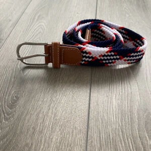 Randigt flätat bälte i blått och vitt - Snyggt flätat bälte med randigt mönster i marinblått, vitt och röda detaljer. Spännet är i silverfärgad metall och ändarna är förstärkta med brunt skinn. Perfekt accessoar för att lyfta din outfit.