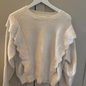 Vit volangtröja från MNG, storlek M - Superfin vit stickad tröja från MNG i storlek M. Tröjan har snygga volangdetaljer längs axlarna och är långärmad med ribbade muddar. Materialmix med ull, polyester och elastan gör den både mjuk och bekväm. Perfekt för dig som gillar en söt och trendig look.