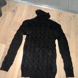 Svart stickad polotröja med kabelmönster - Mysig svart stickad polotröja med klassiskt kabelstickat mönster. Tröjan har hög polokrage och långa ärmar, perfekt för kyliga dagar. Insidan är fodrad med mjukt, ljust tyg för extra komfort. Snygg och enkel att matcha till olika outfits. 9 mån - 2 år. 