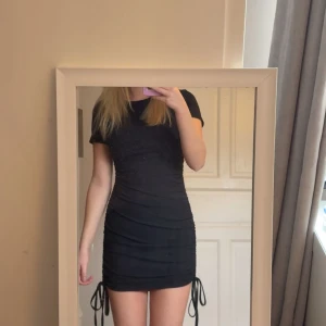 Svart ribbad kortklänning med snörning - Supersnygg svart kortklänning med ribbad struktur och snörning i sidorna. Klänningen har korta ärmar och tight passform som framhäver figuren. Perfekt för dig som gillar en enkel men trendig look. Säljes då den inte kommer till användning. Använd 1 gång
