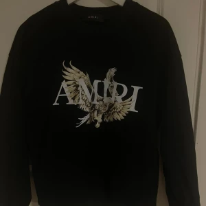 Svart sweatshirt från Amiri med örn - Svart sweatshirt från Amiri med stort tryck av en örn och AMIRI-logga på bröstet. Tröjan har rund hals och långa ärmar, perfekt för dig som vill sticka ut med en cool streetstyle-look.